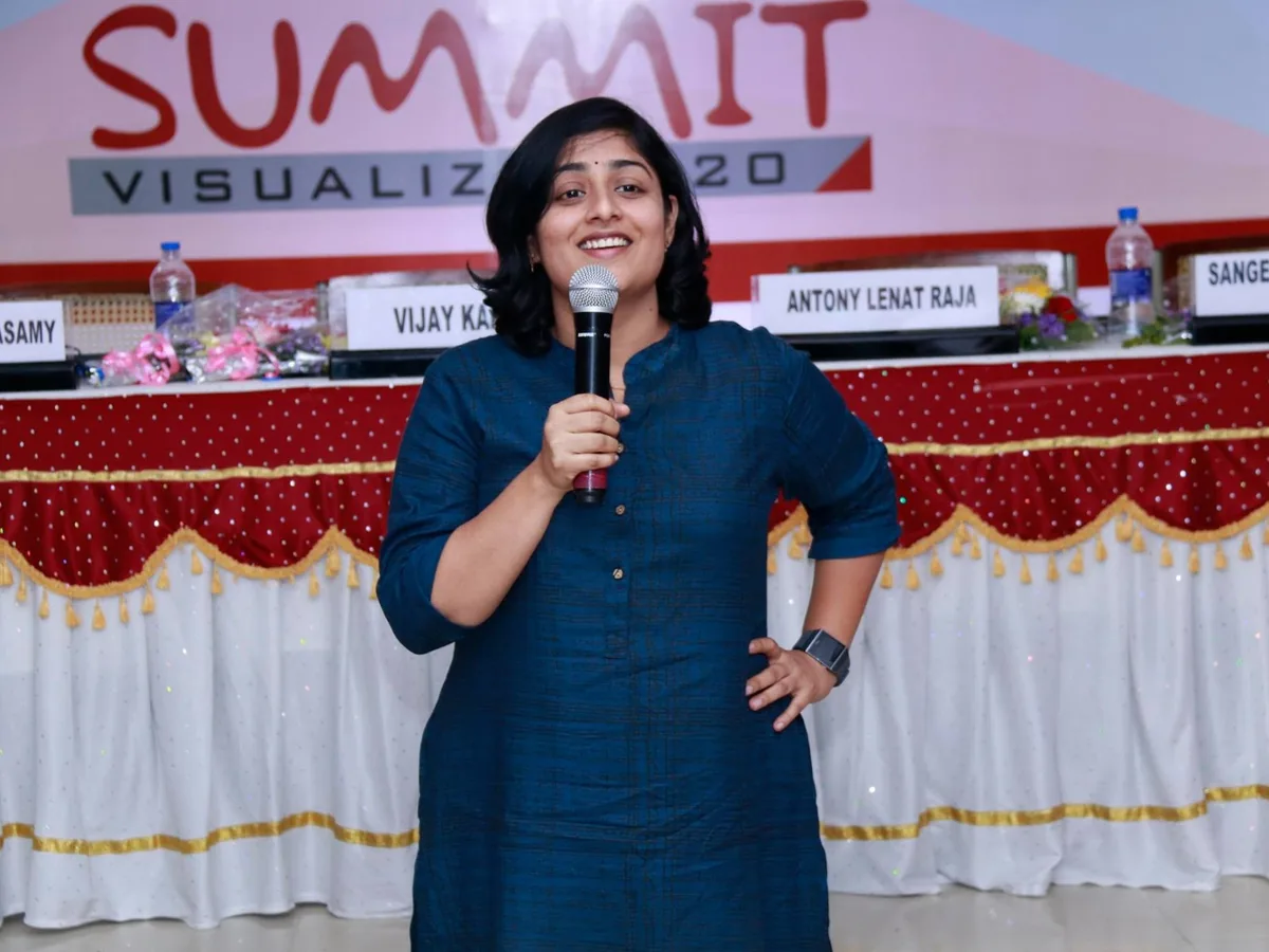 Sangeetha S Abishek speaking with a microphone in front of a summit backdrop and the panel table behind her.