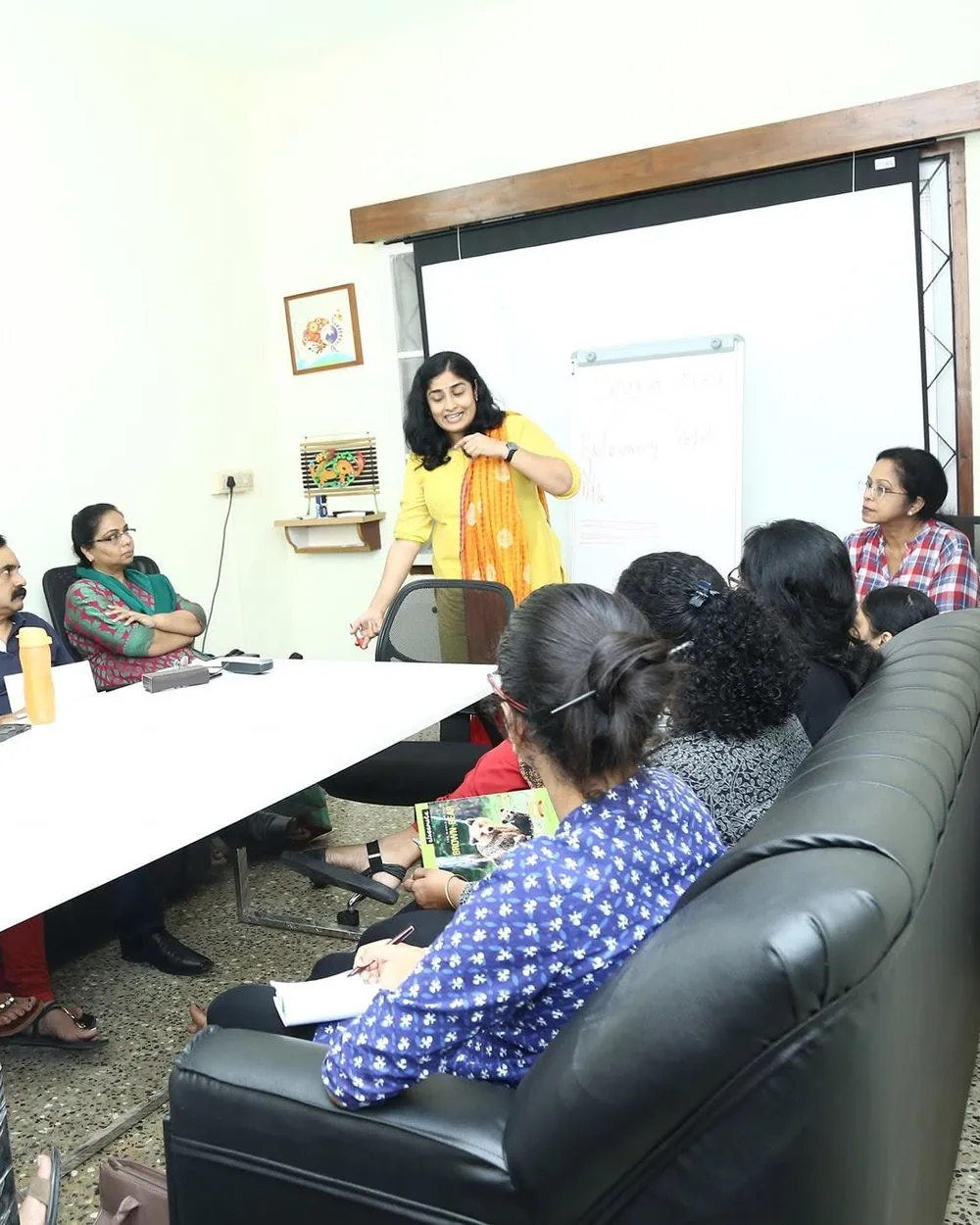 Sangeetha S Abishek working through a plan at a whiteboard with a seated group around a table.